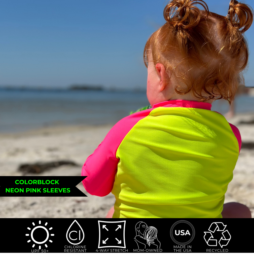 Baby girl sitting on towel on beach sand looking out at ocean wearing a neon yellow rash guard with neon pink sleeves; the bright swimwear stands out against the dull backdrop.